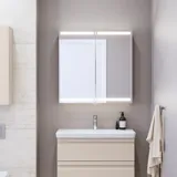Lille badeværelse inspiration med vaskeskab og overskab over toilet i beige specialfarve, elegant spejlskab med integreret lys, de lyse farver og hvide gardin skaber en romantisk og feminin atmosfære.