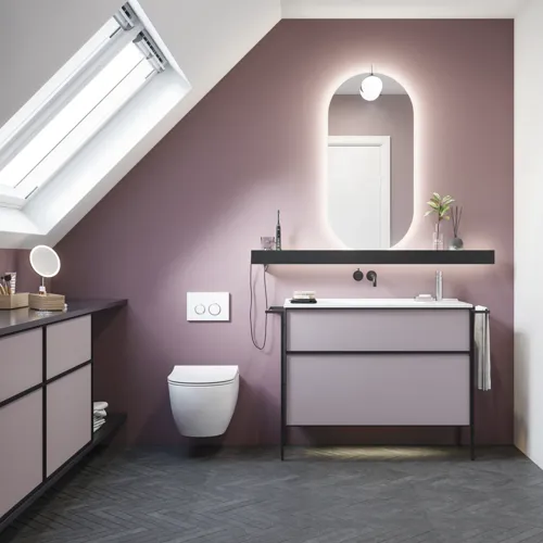 Badeværelse med skrå vægge inspiration, med badeværelsesmøbler i lilla med elegante rammefronter, flot ovalt spejl med lampe, smuk og praktisk hylde, samt lillamalet væg og gråt sildebensgulv, der giver et elegant og moderne udtryk.